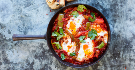 Tomato and pepper shakshuka, topped with four Clarence Court eggs, on a mottled grey background