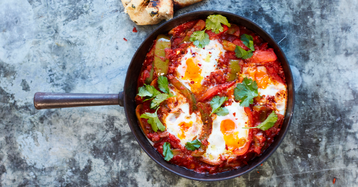 Tomato and pepper shakshuka, topped with four Clarence Court eggs, on a mottled grey background