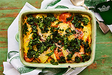 A baking dish of potato and root vegetable gratin, topped with melted cheese and kale, against a white tea towel, decorated with a green egg print.