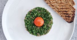 A round portion of steak tartare topped with chopped herbs and an egg yolk rests next to a toasted piece of sourdough bread on a white plate.