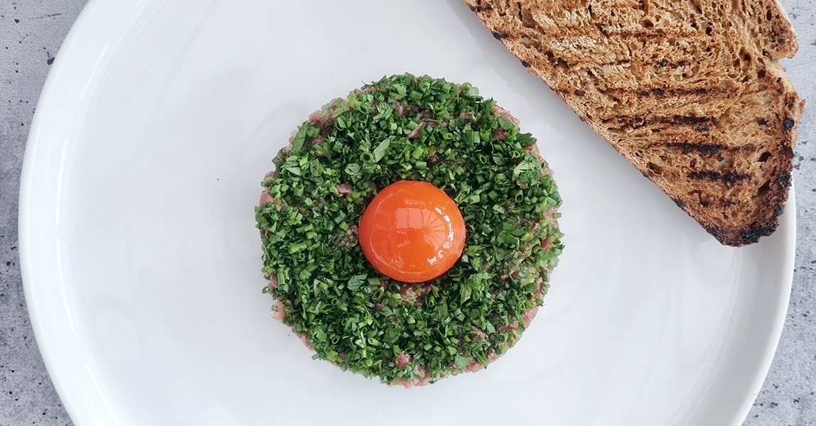 A round portion of steak tartare topped with chopped herbs and an egg yolk rests next to a toasted piece of sourdough bread on a white plate.