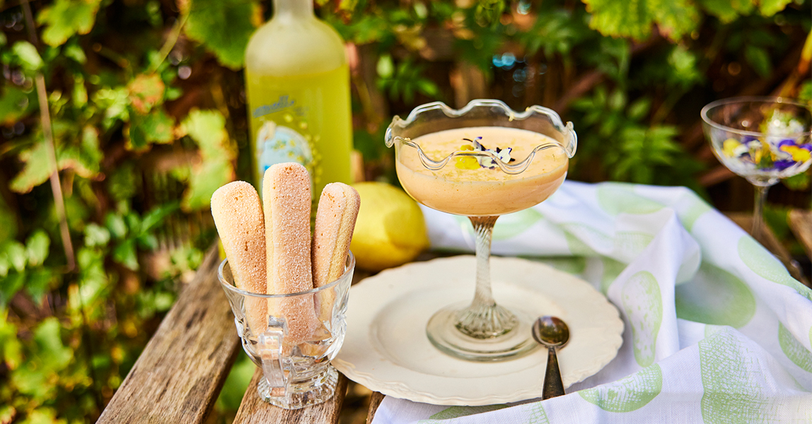 A glass of yellow dessert sits next to a cup of biscuit fingers and a bottle of yellow limoncello