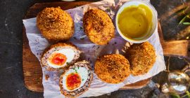 Five Scotch eggs on a wooden board, next to a small bowl of mustard.