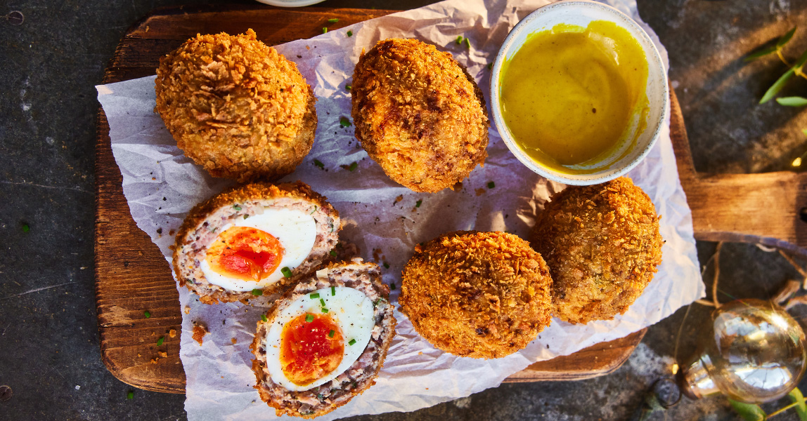 Five Scotch eggs on a wooden board, next to a small bowl of mustard.