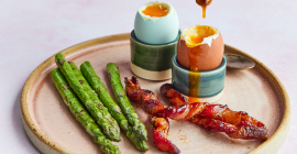 Clarence Court chilli maple bacon soldiers on a plate next to spears of asparagus and two boiled Clarence Court eggs.