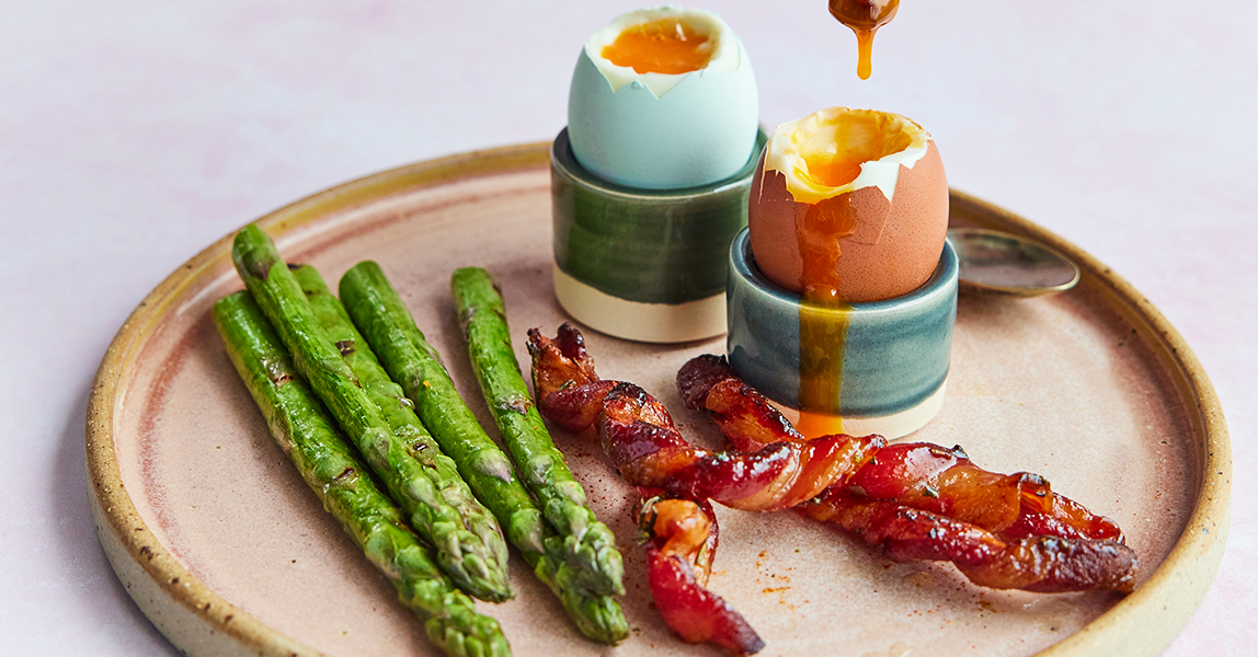 Clarence Court chilli maple bacon soldiers on a plate next to spears of asparagus and two boiled Clarence Court eggs.