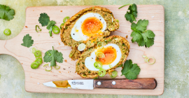Wooden chopping board containing two halves of a Clarence Court Scotch egg.The yolk is gooey and golden, with a firm white and crisp, golden breadcrumb coating. On the board are coriander leaves and a knife with wooden handle.