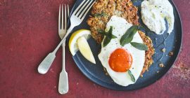 A dinner plate on a red surface with a knife and fork to the side. On the plate are two Jerusalem artichoke fritters, topped with a sage fried Burford Brown egg