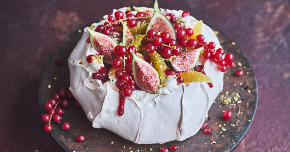 A winter pavlova nest with a topping off winter berries and figs, on a cake stand with a purple backdrop