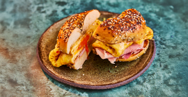 An 'everything bagel' sandwich sits on a stone grey plate on a blue tabletop. inside the sandwich is an egg omelette and ham, which is cut in half so that we can see the central cross-section.