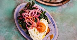 On a purple oval pate sits a slice of cornbread, topped with wilted greens, a fried egg and some thin slices of red onion.
