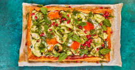 A rectangle of flaky pastry sits on a white tray. Ontop of the pastry sits a medley of cucumber ribbons, spinach, pomegranate seeds, fresh herbs and baked eggs.