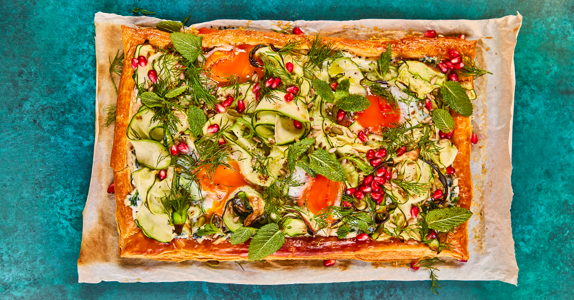 A rectangle of flaky pastry sits on a white tray. Ontop of the pastry sits a medley of cucumber ribbons, spinach, pomegranate seeds, fresh herbs and baked eggs.