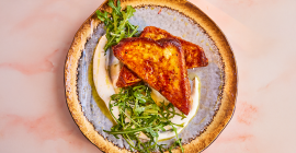 A savoury twist on the classic French toast recipe. On a stone grey plate sits a fried French toast cut in half into triangles. Two bundles of green salad sit to the side of the toast.