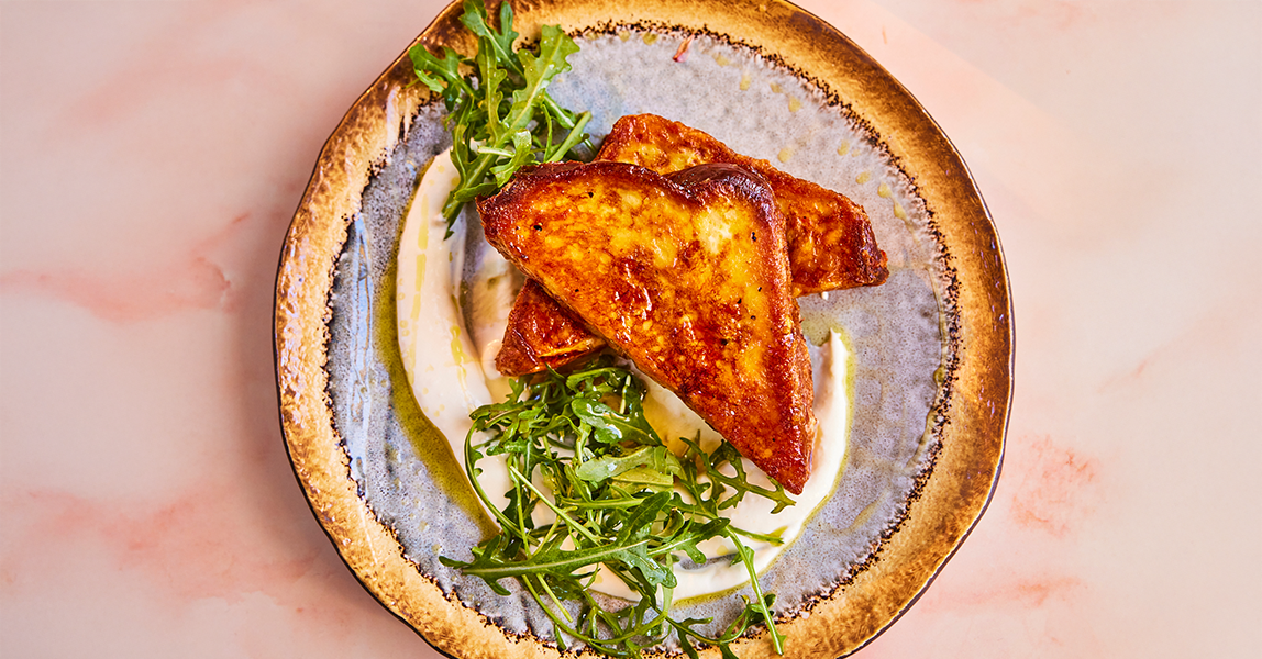 A savoury twist on the classic French toast recipe. On a stone grey plate sits a fried French toast cut in half into triangles. Two bundles of green salad sit to the side of the toast.