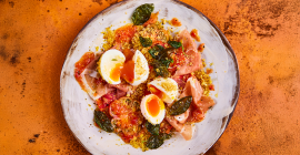 Soft boiled eggs with tomatoes, prosciutto and pangratto sit on a stone grey plate. The soft boiled eggs are cut in half and sit on top of the salad, with the yolk slowly oozing over the top of the tomatoes.