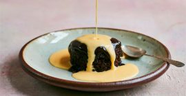 A jug of creme anglaise is being poured from a height, onto a round of sticky toffee pudding.