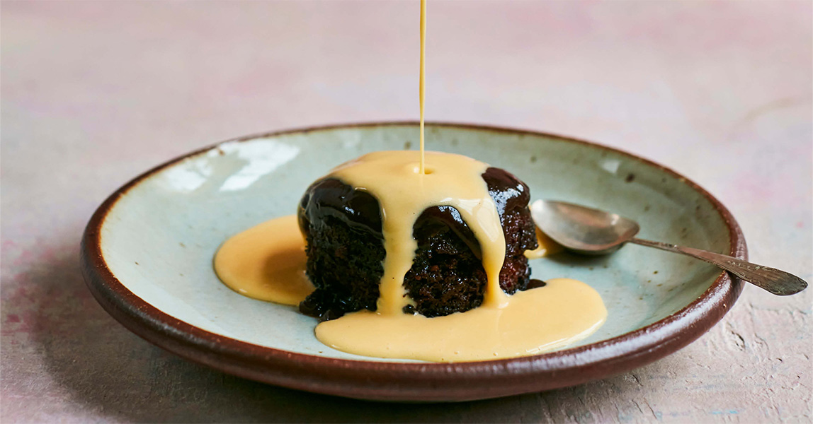 A jug of creme anglaise is being poured from a height, onto a round of sticky toffee pudding.