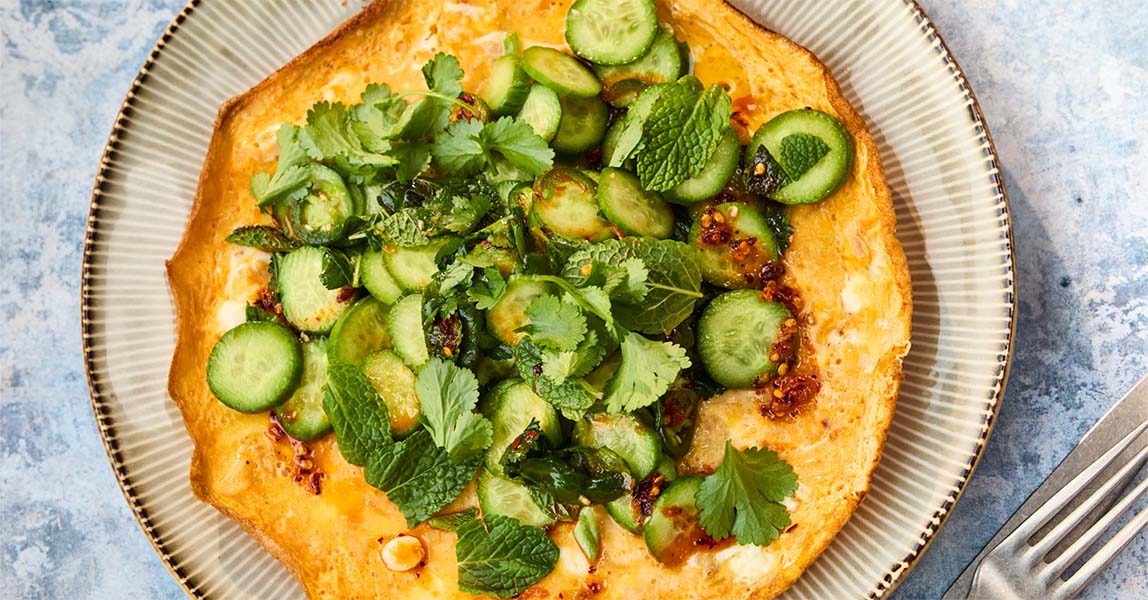 A round crispy rice omelette from Clarence Court sits on a grey plate, which sits on a blue tabletop. Spread across the omelette is slices of cucumber and greens.