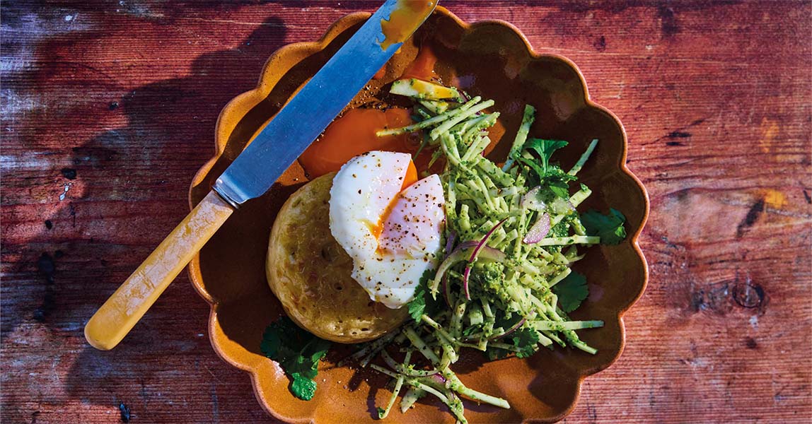 In a scalloped edged terracotta bowl plate, sits a crumpet that has a poached egg on top. The egg has been popped and golden yellow yolk is pouring out. To the right of the crumpet and egg is a handful of julienned celeriac sticks with a green sauce coating them.