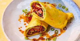 A rice paper egg wrap sits cut in half on a pale grey plate, with a pouring of chilli oil surrounding it.