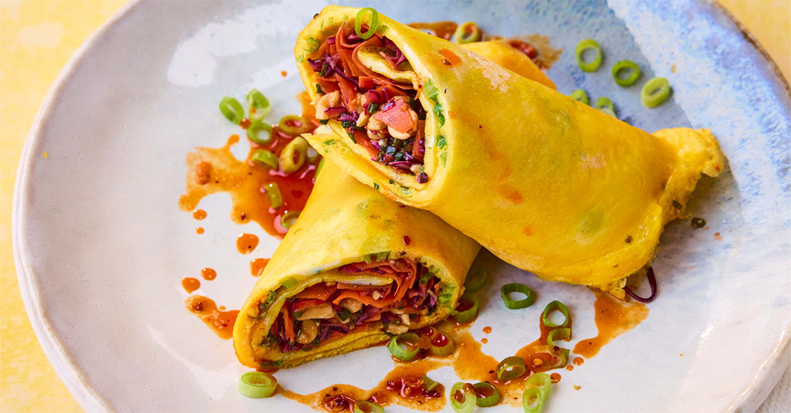 A rice paper egg wrap sits cut in half on a pale grey plate, with a pouring of chilli oil surrounding it.